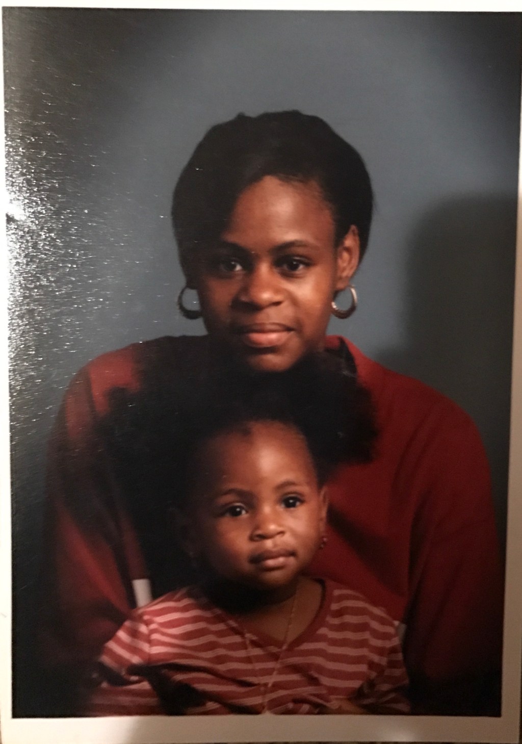 A woman with hoop earrings and a red top sits behind a young girl in a striped shirt, both facing forward against a plain dark background.